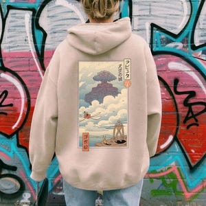 May include: Beige hoodie with a Japanese-style illustration on the back. The design shows a floating castle, figures, and Japanese text. The hoodie has a hood and long sleeves, suitable for everyday wear.