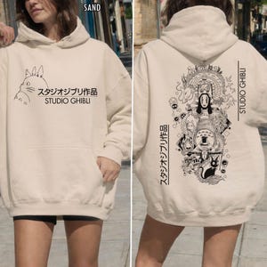 May include: Sand-colored Studio Ghibli hoodie with a front pocket and hood. The front features a Totoro graphic and Japanese text. The back displays a detailed black and white illustration of characters and the words "STUDIO GHIBLI".