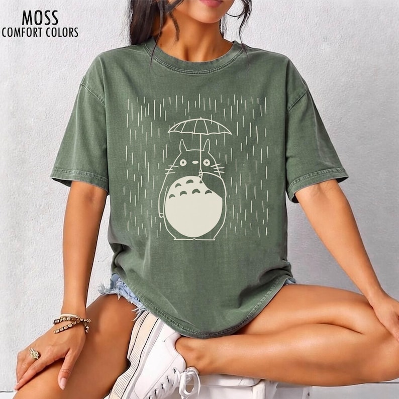 May include: Moss green t-shirt featuring a white illustration of a cartoon character holding an umbrella in the rain. The text "MOSS COMFORT COLORS" is printed in the upper left corner. The shirt has short sleeves and a relaxed fit.