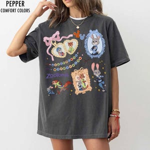 May include: A charcoal gray short-sleeve t-shirt featuring a graphic print of Zootopia characters. The design includes heart-shaped frames, a blue ornate frame, and the text Welcome to Zootopia. The shirt has a relaxed fit and crew neckline.
