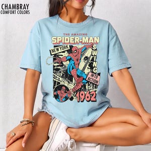 K&ouml;nnte beinhalten: Hellblaues T-Shirt mit einem Vintage-Spider-Man-Motiv. Das Design enth&auml;lt die Schriftz&uuml;ge "THE AMAZING SPIDER-MAN", "NEW YORK", "SWING INTO ACTION" und "1962". Das T-Shirt ist aus weichem Stoff und hat kurze &Auml;rmel.