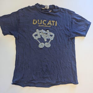 May include: Navy blue t-shirt with distressed edges, featuring the gold text "DUCATI meccanica" above a light grey graphic. The shirt has a vintage look, with visible wear and tear.