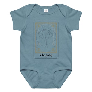 May include: A pale blue baby bodysuit featuring a gold-framed graphic of hands holding baby feet. The text "The baby" is below the graphic. The bodysuit has short sleeves and a snap closure.