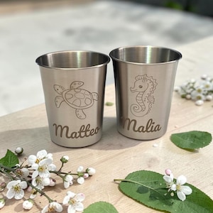 Könnte beinhalten: Zwei Becher aus gebürstetem Edelstahl mit lasergravierten Meeresmotiven. Der linke Becher zeigt eine Meeresschildkröte und den Namen Matteo, der rechte Becher ein Seepferdchen und den Namen Malia, beide in einer klaren, metallischen Optik.