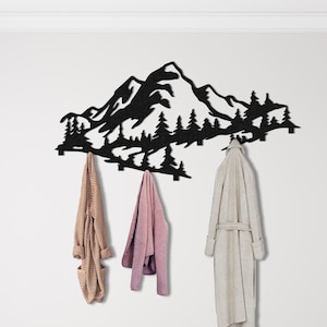 Può includere: Un attaccapanni da parete in metallo nero con un design a silhouette di montagna e foresta. Tre accappatoi beige, rosa e grigio chiaro sono appesi ai ganci. L'attaccapanni è montato su una parete bianca.