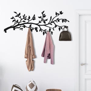 May include: A black metal wall-mounted coat rack in the shape of a tree branch, holding a brown handbag and two garments. Below, two woven baskets and decorative pillows sit on a wooden floor, near a white door.