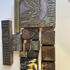 May include: A collection of vintage metal and wood printing blocks featuring various engraved designs, including a large moon graphic, a person holding playing cards, architectural structures, and reversed text elements like "UNO 20MAH".