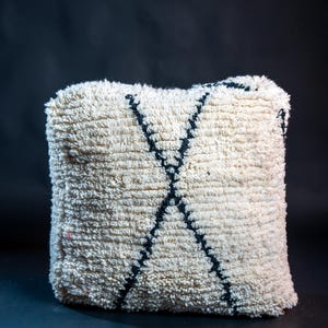 May include: A square, off-white, high-pile shag throw pillow featuring a prominent, dark black, hand-knotted X-shaped pattern centered on the front. The thick, textured wool-like fibers create a dense, soft appearance across the surface.