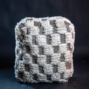 May include: A square throw pillow featuring a checkered pattern of alternating brown woven fabric squares and raised, textured white tufted squares. The pillow has a dense, plush pile on the white sections and a flat, woven texture on the brown sections.