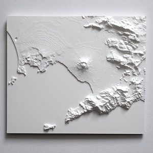 May include: A white, three-dimensional relief map depicting a coastal region with mountains, a volcano, and islands. The map is mounted on a white rectangular surface, showcasing detailed topographical features.