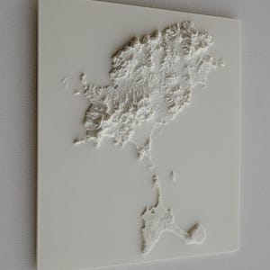 Könnte beinhalten: Eine weiße 3D-topografische Karte einer Insel, die detaillierte Höhenunterschiede zeigt. Die Karte ist auf einer weißen rechteckigen Basis montiert, wobei die Küstenlinie und das Gelände der Insel deutlich definiert sind. Das Relief ist erhaben.