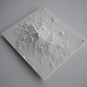 May include: A white, three-dimensional relief map of a mountainous region. The map is square and features detailed topographical features, including peaks, valleys, and ridges. The relief is cast in a solid white material, creating a stark contrast with the surrounding area.