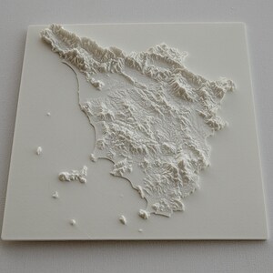 May include: A white, three-dimensional relief map of Tuscany, Italy. The map shows the region's topography with raised features representing mountains, hills, and coastlines. The map is mounted on a square, white base.