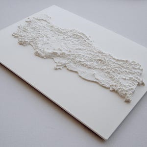 May include: A white relief map of Turkey, showcasing the country's geographical features. The map is mounted on a white rectangular surface, with the relief detail creating shadows and texture. The overall aesthetic is clean and minimalist.