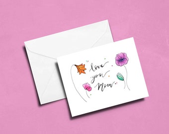May include: A white card with a handwritten message that says "love you Mom" with colourful flowers drawn around the text.