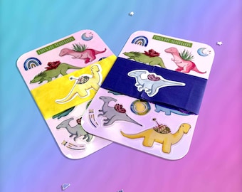 May include: Two sheets of colourful dinosaur stickers with the text "Dinos of Succulents" on each sheet. The stickers feature dinosaurs with succulents growing on their backs. The stickers are on a pink and blue gradient background.