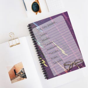 Puede incluir: Una libreta de espiral morada con una hoja de cubierta de plástico transparente. La hoja de cubierta tiene un diseño abstracto colorido y el texto "The Daily Schedule" escrito en ella. La libreta tiene páginas rayadas y un par de gafas están sobre la cubierta.