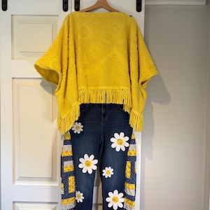 May include: A yellow poncho with fringe detail hangs above a pair of dark wash denim jeans. The jeans feature large white daisy appliqués and patchwork accents with yellow and navy blue patterns. The outfit is displayed on a white door.