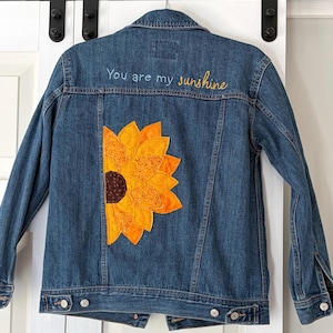 May include: A blue denim jacket with a large, appliqued sunflower design on the back. The sunflower is made of orange and yellow fabric with a brown center. The words "You are my sunshine" are embroidered in yellow thread above the sunflower.