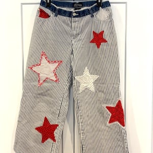 May include: Wide-leg, striped pants with a denim waistband. The pants feature vertical black and white stripes and are adorned with red and white star appliqués. The pants are hanging on a black hanger against a white door.