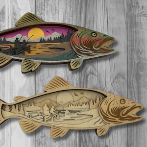 May include: Two wooden fish-shaped wall art pieces. The top fish has a colourful interior scene of a lake with a sunset, mountains, and two people fishing. The bottom fish has a similar scene in a natural wood tone.