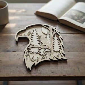 Może przedstawiać: Wielowarstwowe drewniane dzieło sztuki w kształcie głowy orła. Projekt przedstawia szczegółową scenę leśną z drzewami, orłem i pełnią księżyca. Grafika jest wyświetlana na drewnianym stole.