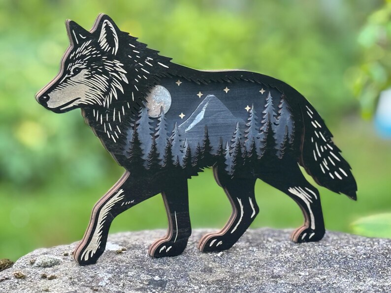 May include: A wooden wolf figurine with a detailed design. The wolf is black with a light beige snout and paws. The interior features a mountain scene with trees, a moon, and stars. The figurine is approximately 10 inches long.