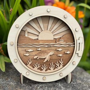 May include: A round, wooden nautical-themed wall art piece. The artwork features a layered design with a lighthouse, sun, boat, turtle, and fish. The outer frame is light-colored, with a darker, natural wood interior. It is displayed on a stand.