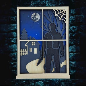 May include: Wooden layered art piece depicting a spooky scene through a window. The artwork features a silhouette of a figure holding a knife, a house, a full moon, and a spiderweb. The color palette includes blue, black, and natural wood tones.
