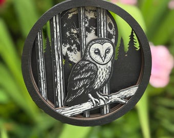 Op de afbeelding: Een ronde, zwarte houten ornament met een gedetailleerde uil op een tak. De uil staat tegen een achtergrond van een volle maan en een boslandschap. Het ornament hangt aan een zwart koord en is omgeven door groene bladeren.