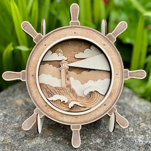 Può includere: Un timone di nave in legno con una scena di faro all'interno. Il timone è marrone chiaro con accenti marroni più scuri. Il faro è bianco, sullo sfondo di nuvole, onde e luna piena. Il pezzo è esposto su una roccia grigia.