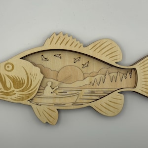 Op de afbeelding: Houten wandkunst in de vorm van een vis met een gelaagd ontwerp. De binnenkant toont een sereen landschap met een visser in een boot, vogels, bomen en een ondergaande zon. De vis is lichtbruin met gedetailleerde vinnen en schubben.