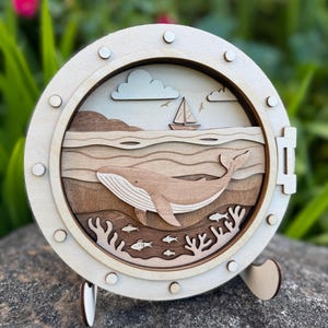 May include: Wooden nautical-themed wall art featuring a whale, sailboat, and ocean scene. The artwork is layered with various shades of brown and tan, set within a round, porthole-like frame. The piece is mounted on a stand.