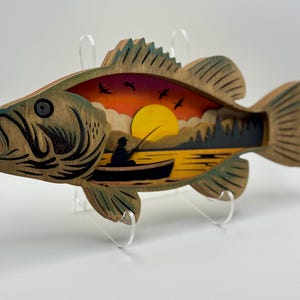 Op de afbeelding: Houten wandkunst in de vorm van een baarsvis met een gelaagd ontwerp. De binnensc&egrave;ne toont een zonsondergang boven een meer met een visser in een boot. De vis heeft een natuurlijke houtnerf afwerking met groene accenten.