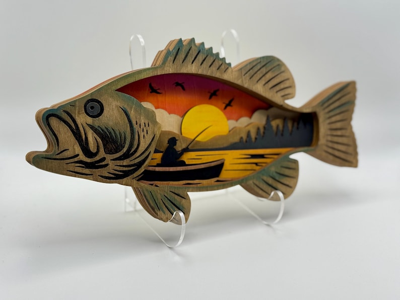 Op de afbeelding: Houten wandkunst in de vorm van een baarsvis met een gelaagd ontwerp. De binnensc&egrave;ne toont een zonsondergang boven een meer met een visser in een boot. De vis heeft een natuurlijke houtnerf afwerking met groene accenten.