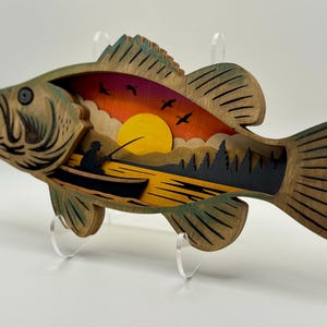 Op de afbeelding: Houten kunstwerk in de vorm van een vis met een gelaagd ontwerp dat een zonsondergangsc&egrave;ne uitbeeldt. Het kunstwerk toont een visser in een boot, bomen, vogels en een gele zon tegen een kleurrijke lucht. De vis is geschilderd in bruin- en groentinten.
