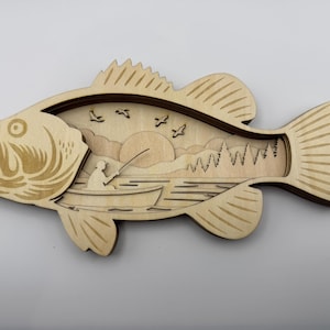 Op de afbeelding: Houten wandkunst van een basvis met een gelaagd ontwerp. De binnenkant toont een visser in een boot, een zonsondergang, bomen en vogels. De vis is lichtbruin met donkerbruine accenten.