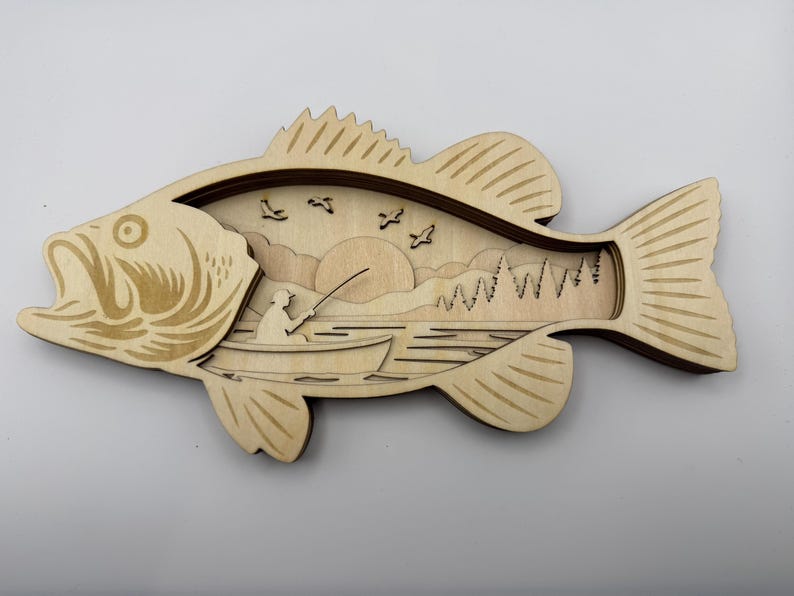 Op de afbeelding: Houten wandkunst van een basvis met een gelaagd ontwerp. De binnenkant toont een visser in een boot, een zonsondergang, bomen en vogels. De vis is lichtbruin met donkerbruine accenten.