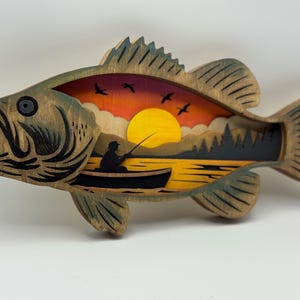 Op de afbeelding: Houten kunstwerk in de vorm van een vis met een gelaagd ontwerp dat een zonsondergangsc&egrave;ne uitbeeldt. Het kunstwerk toont een visser in een boot, bomen, vogels en een gele zon. De vis heeft een natuurlijke houtnerf afwerking met groene accenten.