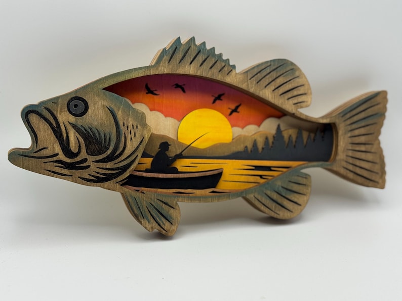 Op de afbeelding: Houten kunstwerk in de vorm van een vis met een gelaagd ontwerp dat een zonsondergangsc&egrave;ne uitbeeldt. Het kunstwerk toont een visser in een boot, bomen, vogels en een gele zon. De vis heeft een natuurlijke houtnerf afwerking met groene accenten.