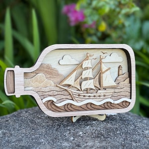 May include: A wooden ship in a bottle art piece. The multi-layered design features a sailing ship, waves, clouds, and a lighthouse. The artwork is displayed on a wooden stand, showcasing the intricate details of the laser-cut design.