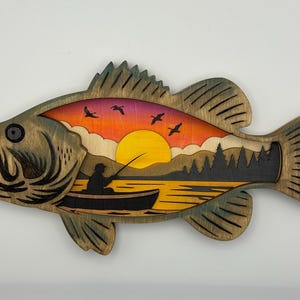 Op de afbeelding: Houten wandkunst van een basvis met een gelaagd ontwerp. De binnenkant toont een zonsondergangsc&egrave;ne met een visser in een boot, vogels en bomen. De vis is geschilderd in groen- en bruintinten.