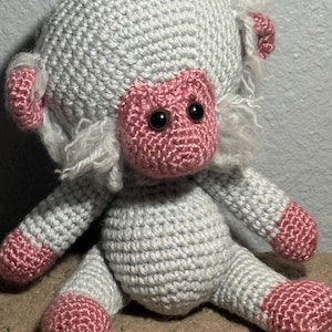 May include: A crocheted stuffed animal resembling a monkey with light gray yarn for the body and head, and pink yarn for the face, ears, hands, and feet. The toy features dark brown plastic eyes and tufts of fuzzy white yarn around the ears.
