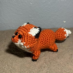 May include: A small, handcrafted crochet fox toy featuring vibrant orange yarn, white accents on the face and tail tip, and black yarn details on the ears, feet, and nose. The fox has two round, glossy black plastic eyes and a soft, stuffed texture.