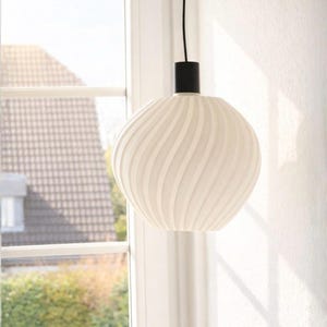 Könnte beinhalten: Eine weiße, kugelförmige Pendelleuchte mit einem wirbelnden, gerippten Design. Die Leuchte hängt an einer schwarzen zylindrischen Basis und einem schwarzen Kabel. Das Licht befindet sich vor einem Fenster.
