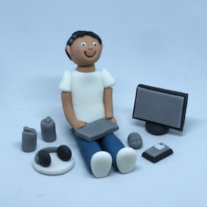 May include: A figurine of a person sitting with a computer, keyboard, mouse, and headphones. The person is wearing a white shirt and blue jeans. There are two grey cans next to the person.