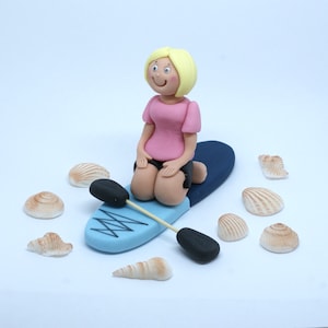 May include: A blonde figurine of a person sitting on a blue and white surfboard with a paddle. The figurine is wearing a pink shirt and black shorts. There are several seashells scattered around the figurine.