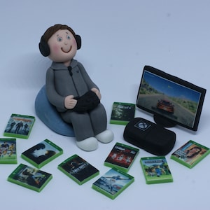 May include: A figurine of a person wearing a grey hoodie and headphones, sitting on a blue beanbag chair, holding a video game controller. The person is in front of a black computer monitor displaying a video game. There are several video game cases scattered around the figurine.