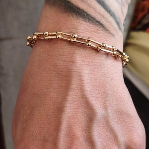 Könnte beinhalten: Ein goldfarbenes Armband mit Kettenglied-Design. Das Armband hat eine Reihe von rechteckigen Gliedern, die durch kleine, runde Kugeln verbunden sind. Der Schmuck wird am Handgelenk einer Person getragen.