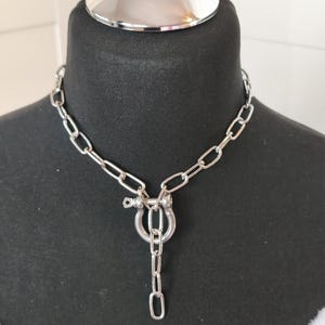 Könnte beinhalten: Silberfarbene Halskette mit einem großen, offenmaschigen Design. Die Halskette hat einen markanten U-förmigen Anhänger mit einer herabhängenden Kette und einem sicheren Verschluss. Der Schmuck wird auf einer schwarzen Schaufensterpuppe präsentiert.